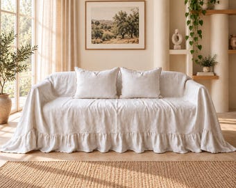 May include: A cream-coloured sofa covered with a long, draped fabric slipcover featuring a ruffled bottom edge. Two matching square throw pillows are placed on the seat. The fabric has a visible, textured weave, and the entire piece is presented in a neutral, light-toned setting.
