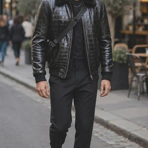 May include: A black jacket with a textured crocodile-skin pattern, a black faux fur collar, and ribbed cuffs. The jacket is worn over a plain black t-shirt and paired with black trousers and black leather boots. A black crossbody bag strap is visible.