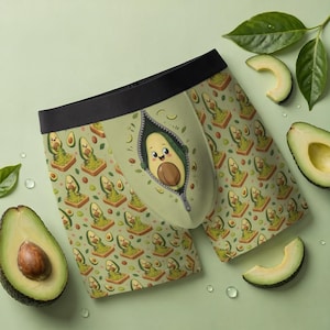 Op de afbeelding: Boxershorts met een lichtgroene basis en een herhalend patroon van cartoon avocado's op toast. Een centrale afbeelding toont een lachend avocado-personage dat uit een ritsopening tevoorschijn komt. Het kledingstuk heeft een zwarte elastische tailleband.
