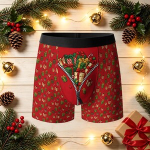 May include: Red boxer briefs featuring a repeating pattern of small Christmas gifts and candy canes. The centre panel displays a larger graphic of stacked presents behind a partially open zipper design. The garment has a solid black waistband.