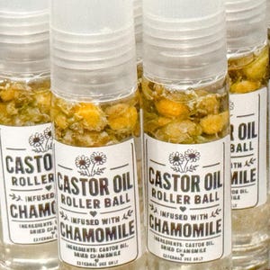 Può includere: Flaconi in vetro trasparente con applicatore a sfera, riempiti con olio di ricino e fiori di camomilla gialli essiccati. Ogni flacone presenta un'etichetta bianca con il testo CASTOR OIL ROLLER BALL INFUSED WITH CHAMOMILE, ingredienti e un'illustrazione floreale.