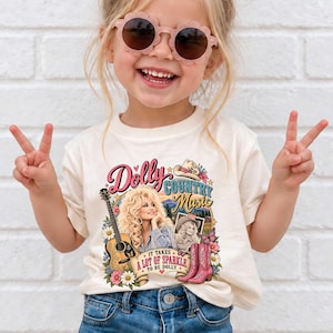 Puede incluir: Una camiseta color crema con un estampado gráfico de una mujer rubia, una guitarra acústica, un sombrero de vaquero y botas rosas. El texto dice Dolly Country Music y It Takes A Lot Of Sparkle To Be Dolly, rodeado de motivos florales.