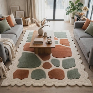 May include: A rectangular area rug with a wavy scalloped edge featuring an abstract pattern of irregular, rounded shapes in shades of sage green and terracotta orange set against a cream-colored background, covering the floor between two sofas.