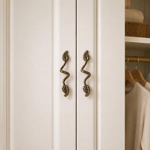 May include: Two antique brass cabinet handles with a winding, vine-like shape featuring leaf details at the top and bottom. These metal pulls are mounted vertically on the white, panelled doors of a wardrobe, providing a decorative accent.