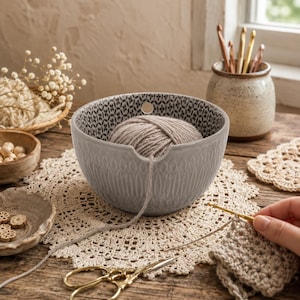 Peut inclure: Un bol à laine en céramique gris clair avec un extérieur texturé et un intérieur à motifs noirs et blancs. Le bol présente un trou circulaire et une encoche en forme de U pour le fil, contenant une pelote de laine beige qui passe par l'encoche.
