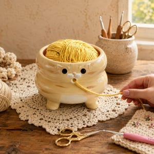 May include: A ceramic yarn bowl shaped like a light orange tabby cat with a glossy finish. The bowl features a small hole near the cat's mouth for feeding yarn, and it is filled with a ball of bright yellow yarn. The cat has painted eyes.