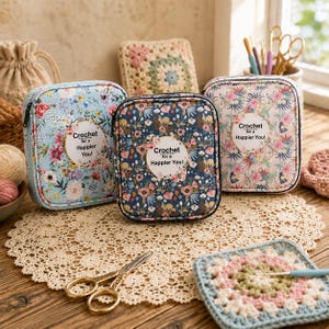 Può includere: Tre astucci rettangolari con cerniera e motivi floreali in azzurro, blu scuro e rosa chiaro. Ogni astuccio presenta un'etichetta bianca circolare con il testo "Crochet for a Happier You!" centrato sulla parte anteriore.