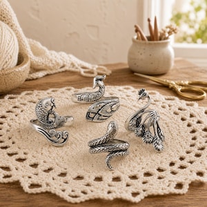 May include: A collection of four silver-toned metal rings featuring detailed animal designs. The set includes a koi fish ring, a coiled snake ring, and two distinct peacock rings with intricate feather patterns, all displayed on a cream-colored lace mat.
