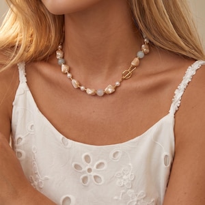 May include: A necklace featuring irregular peach-toned beads, small white pearls, and smooth light blue spherical beads, accented with tiny gold-toned spacers. The piece is secured with a gold-toned toggle clasp positioned at the front.