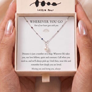 Pode incluir: Um colar de corrente prateada com pequenas pedras coloridas incrustadas e um pingente de pérola branca, apresentado numa caixa de cor creme. O cartão no interior diz: WHEREVER YOU GO, Part of our heart goes with you. Distance is just a number.