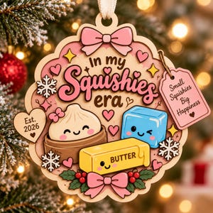 以下が含まれることがあります： In my Squishies eraとSmall Squishies Big Happinessの文字が入った木製オーナメント。蒸し器に入った餃子、青いキューブ、顔の描かれた黄色いバターのイラストが描かれ、ピンクのリボン、ハート、雪の結晶で装飾されています。