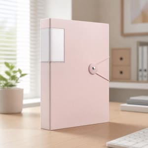 Puede incluir: Una carpeta vertical de color rosa claro que presenta un portaetiquetas rectangular blanco en el lomo y un cierre de botón circular con un cordón elástico que envuelve la cubierta frontal para asegurar el contenido dentro del archivador.