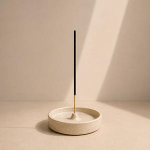 Peut inclure: Un porte-encens en céramique blanc cassé de forme circulaire avec une texture mouchetée et un cône central surélevé doté d'un petit trou. Un bâton d'encens noir, fin et unique, est inséré verticalement au centre du support.