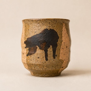 Pode incluir: Uma chávena cilíndrica de cerâmica com uma superfície texturizada em tons de castanho e bege salpicado. O exterior apresenta uma forma abstrata irregular de cor castanho escuro na parte frontal. O rebordo é ligeiramente alargado.