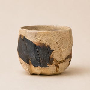 Może przedstawiać: Mały, okrągły kubek ceramiczny o teksturowanej, beżowej, piaskowej powierzchni. Zewnętrzna strona posiada wyraźną, nieregularną plamę matowego czarnego szkliwa z widocznymi pionowymi rowkami i subtelnymi brązowymi akcentami na krawędziach.