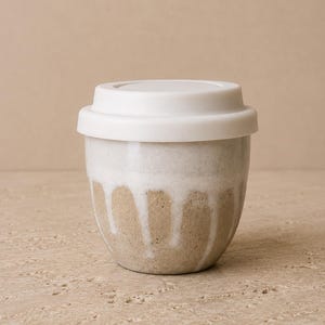 Könnte beinhalten: Ein Keramik-Reisebecher mit einem weißen Silikondeckel. Der Becher hat eine gesprenkelte beige Basis mit einer weißen Glasur, die vom Rand herabläuft und ein strukturiertes Aussehen erzeugt. Das Design ist schlicht und funktional, mit einer runden Form.