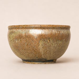 May include: A round ceramic bowl with a speckled, mottled glaze featuring earthy tones of brown, tan, and muted green. The bowl has a smooth, glossy finish with a slightly flared rim and a rounded base, showcasing a variegated texture throughout.