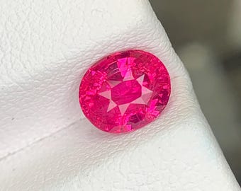 Puede incluir: Una gema de rubí de corte ovalado vibrante con un tono rojo rosado intenso y una superficie multifacetada, que descansa sobre una tarjeta de presentación blanca texturizada. La piedra muestra reflejos internos claros y un acabado pulido.