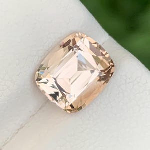 May include: A cushion-cut gemstone with a pale peach or champagne hue. The stone features a faceted surface that reflects light, creating a clear, transparent appearance with sharp edges and a symmetrical, rectangular shape with rounded corners.