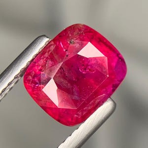 以下が含まれることがあります： 鮮やかで飽和した赤色と内部のインクルージョンが目に見える、クッションカットのルビーの宝石。宝石は光を反射するファセット加工された表面を持ち、深い深紅の色合いと磨かれた半透明の外観を特徴としています。