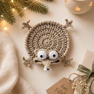 May include: A round, beige crochet coaster shaped like a cartoon creature with four star-like limbs, two large white eyes with black pupils, and a small snout featuring a single white tooth. The coaster is crafted from textured yarn.