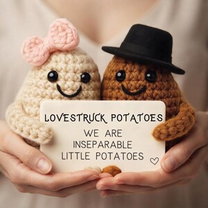 Pode incluir: Duas figuras de batata em crochê, uma cor creme com um laço rosa e uma marrom com um chapéu preto, segurando um cartão branco que diz: LOVESTRUCK POTATOES WE ARE INSEPARABLE LITTLE POTATOES com um pequeno símbolo de coração no canto.