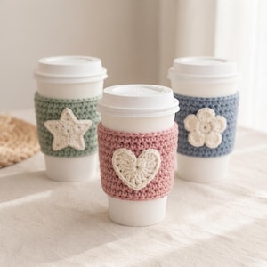 Puede incluir: Tres vasos de café desechables blancos con fundas de ganchillo texturizadas. El vaso izquierdo tiene una funda verde salvia con una estrella crema, el central una funda rosa empolvado con un corazón crema y el derecho una funda azul con una flor crema.