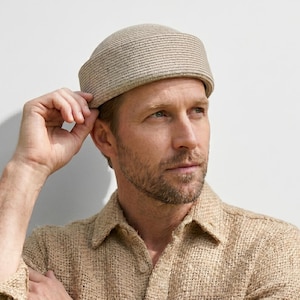 Peut inclure: Une casquette beige sans bordure à texture côtelée, portée avec une chemise à manches longues boutonnée en tissu texturé beige et une montre-bracelet dotée d'un bracelet en cuir marron et d'un cadran blanc, dans un style neutre.
