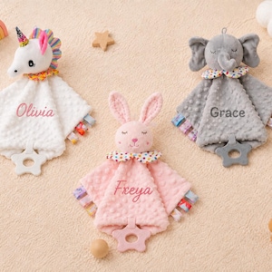 May include: Three plush security blankets featuring a white unicorn with the name Olivia, a pink rabbit with the name Freya, and a gray elephant with the name Grace. Each blanket has a dimpled texture, colorful ribbon tags, and a star-shaped teether.