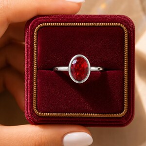 May include: A silver ring featuring a central oval-cut red gemstone set in a smooth bezel. The ring is presented inside a square, deep red velvet jewellery box with gold-toned trim along the edges of the lid and the base of the container.