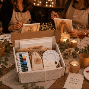 May include: An artist kit in a cardboard box containing a set of colourful paint tubes, a white plastic palette, a small beige drawstring bag labelled The Artist Kit, and wooden easel parts. A card reads Sip. Paint. Mingle. Repeat. and Dabble & Mingle.