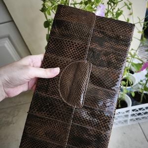 May include: A rectangular clutch bag featuring a dark brown snakeskin pattern with a glossy finish. The bag has a structured design with a central flap closure that is integrated into the front panel, showcasing a consistent scale texture throughout.