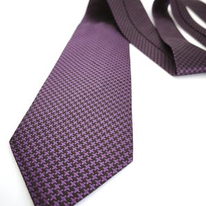 Peut inclure: Une cravate avec un motif pied-de-poule violet et noir. La cravate est disposée en diagonale, montrant l'extrémité pointue et le tissu plié de la bande de cou. La texture tissée du tissu est visible sur toute la longueur de la cravate.