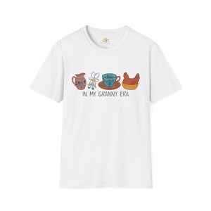 May include: A white short-sleeved t-shirt featuring a graphic print of a floral pitcher, two spoons tied with a ribbon, a teacup on a saucer, and a hen in a basket. Below the illustrations, the text reads IN MY GRANNY ERA in black lettering.