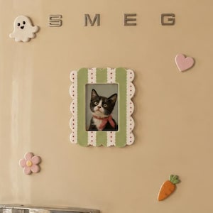May include: A cream-colored SMEG refrigerator featuring a rectangular scalloped picture frame with a cat photo, surrounded by five decorative magnets: a white ghost, a pink heart, a pink flower, and an orange carrot with green leaves.