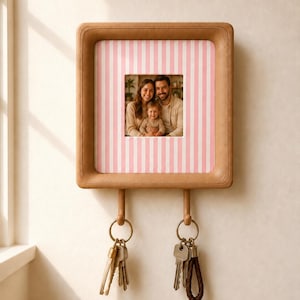 May include: A square, tan-colored wall-mounted key holder featuring a central photo frame displaying a family portrait. The frame is surrounded by a pink and white vertical striped border. Two wooden pegs extend from the bottom, each holding a metal keyring.