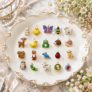 Può includere: Una collezione di diciannove piccoli ciondoli in smalto colorato raffiguranti vari animali e motivi naturali, tra cui un coniglio, un pulcino, una farfalla, un'ape, una pecora, un cervo, un'anatra, una rana, un riccio, una lumaca, uno scoiattolo, una volpe, un uccellino blu, una coccinella, un cane, un gufo, un bruco, una capra e una fragola.
