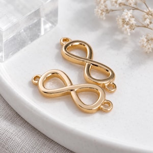 May include: Two gold-toned metal infinity symbol connectors, each featuring a smooth, polished finish and two small circular loops on the ends for attachment. These jewellery components are shaped as horizontal figure-eight loops with open centres.