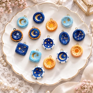 May include: A collection of small, circular and square jewelry charms featuring celestial motifs including gold-painted suns, moons, and star constellations. The charms are rendered in shades of deep blue, light blue, and orange, each with a metal loop.