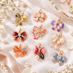 May include: A collection of nine decorative bow-shaped pendants featuring enamel finishes in various colors including red, pink, orange, purple, blue, and cream. Each pendant includes small metallic loops, embedded crystals, and floral accents.