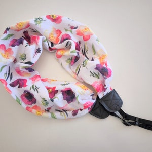 May include: A white camera strap with a floral print featuring pink, red, and purple flowers. The strap has a black adjustable buckle.
