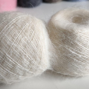 May include: Two fluffy, off-white yarn balls. The yarn appears soft and delicate, with a slightly fuzzy texture. The balls are round and sit close together, suggesting they are part of a larger collection of craft supplies.