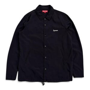 Könnte beinhalten: Eine schwarze langärmelige Coach-Jacke mit spitzem Kragen, einer Druckknopfleiste auf der Vorderseite und zwei Seitentaschen. Das Wort Supreme ist in weißer Schreibschrift auf der linken Brust gestickt und ein rotes Label befindet sich am Kragen.