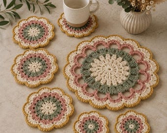 May include: A set of round, crocheted doilies and coasters featuring a floral-inspired pattern in cream, sage green, dusty rose, and mustard yellow. The set includes one large circular mat and six smaller coasters, all with scalloped edges.