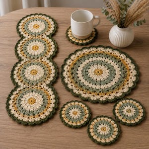 May include: A set of crocheted table linens featuring a circular floral pattern in shades of sage green, mustard yellow, and cream. The collection includes a long runner, one large placemat, and four small coasters, all with scalloped edges.