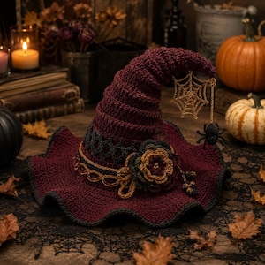 May include: A deep burgundy crocheted witch hat featuring a wide, wavy brim and a pointed crown. The hat is adorned with a dark grey crocheted band, a multi-layered flower accent, and a dangling gold-toned spiderweb charm with a small black spider.