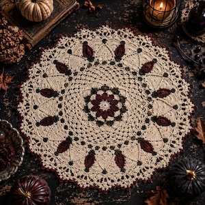 May include: A round, cream-colored crochet doily featuring a central floral motif in dark red and olive green. The intricate lace pattern radiates outward with repeating dark red petal-like shapes and olive green accents, finished with a scalloped edge.