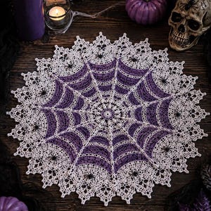 May include: A circular crochet doily featuring a spider web pattern in white and purple yarn. The intricate design includes small black spider embellishments attached to the web structure, with a scalloped, lace-like border edge.