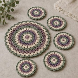 Può includere: Un set composto da un centrino grande e cinque piccoli all'uncinetto, caratterizzati da un motivo circolare concentrico nei toni del verde salvia, crema e viola tenue. Ogni pezzo presenta un bordo smerlato e una trama in filato.