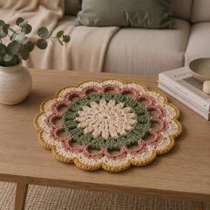 May include: A round, crocheted doily featuring a central cream-colored flower pattern, surrounded by a ring of sage green, a layer of dusty rose, and an outer scalloped border in mustard yellow, creating a layered, circular textile design.